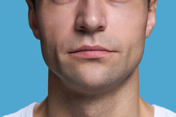 Caucasian young adult man showing lower half of face with neutral expression against blue background, lips closed, stubble visible, wearing casual shirt