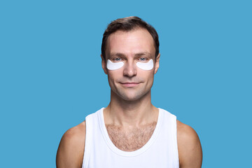 Portrait of Caucasian middle aged man looking at camera wearing under eye patches smiling slightly standing against blue background showing self care routine