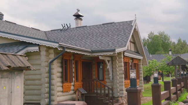 Beautiful traditional wooden house with a shingle roof and intricately carved window frames, showcasing classic slavic architecture in a rural countryside setting with a green lawn and wooden fence