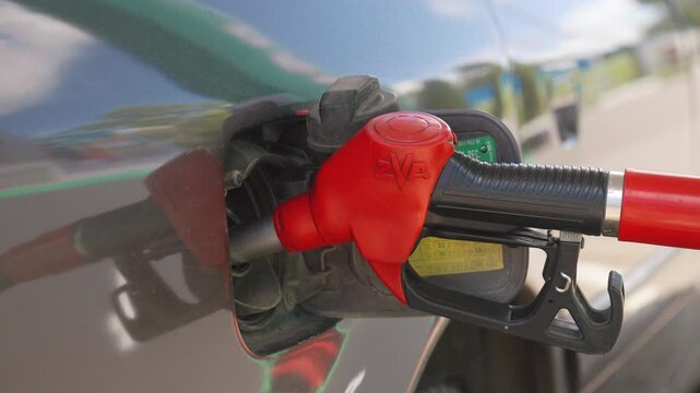 Close-up of a red fuel pump gun nozzle in a dark tank of a car as maintenance personnel refuel the car with gasoline at a gas station, illustrating rising fuel prices, energy crisis and transportation