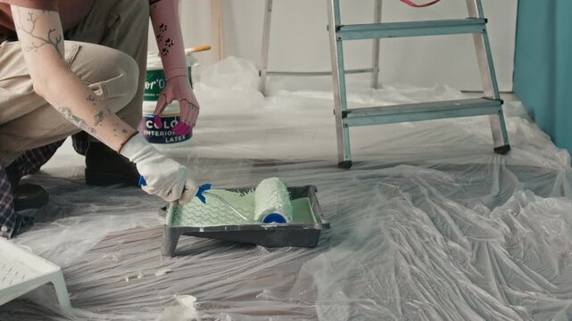 Midsection view of unrecognisable girl with bionic arm using roller brush to apply light green paint poured into plastic tray, with step ladder and paint buckets in background