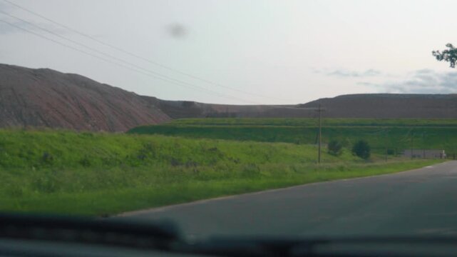 Personal perspective driving along a quiet rural road, passing a large industrial slag heap from a mining operation under a bright sky, illustrating travel through a modified landscape