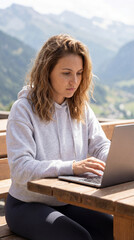 Concentrated young Swiss woman typing on her laptop on a sunny mountain terrace with a scenic alpine view in the background. Modern remote work and flexible workplace concept in nature.
