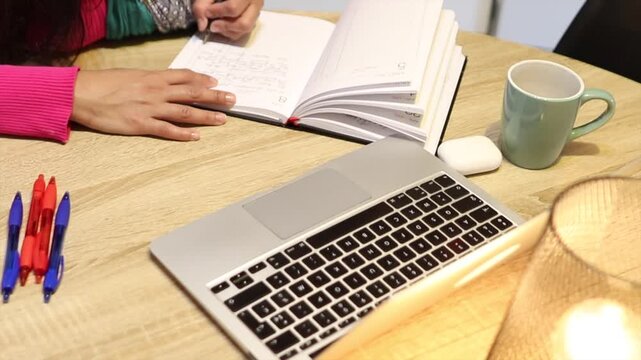 Woman's hands jotting down tasks in a planner using a pen. A student or working woman keeping her day in order, seated at a wooden table with a laptop, a lamp, and a cup of coffee beside her
