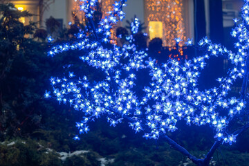 Tree decorated with blue LED flower lights at night. Festive illuminated tree with glowing blue floral lights and warm background in urban celebration setting