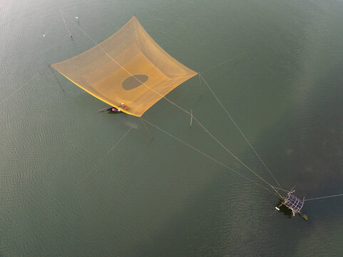 Aerial view of a square fishing net cast over calm waters, creating a striking geometric pattern against the dark water, Hoi An, Quang Nam, Vietnam.