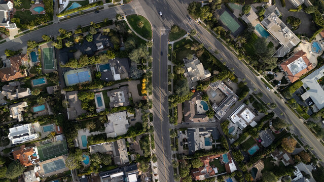 Aerial view of sun-drenched streets slicing through manicured estates, pools glittering like jewels amidst lush greenery, shadows stretching long across Beverly Hills, California, United States.