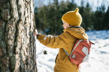 Child in mustard jacket and yellow knit hat stands in snowy forest with a backpack, smiling into...