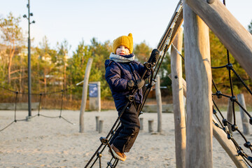 Child in winter clothes on a rope structure on a sandy seaside playground lit by low sun. A bright, brisk moment of joy and movement against the calm Baltic backdrop.
