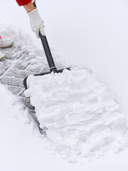 A person wearing white gloves using a black snow shovel to clear fresh white snow from a paved outdoor area. Shallow depth of field.