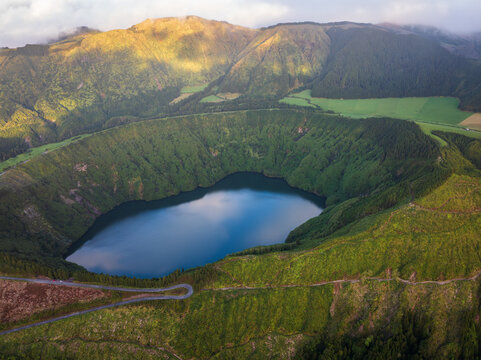 Aerial view of the serene blue lake nestled within the verdant crater, framed by rolling hills and distant fields, Sao Miguel, Azores, Portugal.