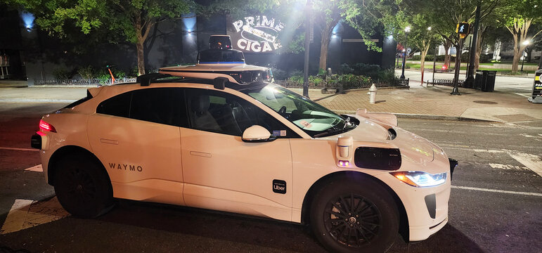 A sleek white autonomous vehicle branded with Waymo is captured in a clean side profile against an urban backdrop at night in Atlanta. The car features prominent rooftop lidar sensors 