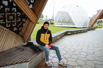 man sitting on bench near dome remote worker vibe yellow-black jacket beanie backpack casual jeans and sneakers open plaza pathway futuristic geodesic structure cloudy sky reflective mood ready