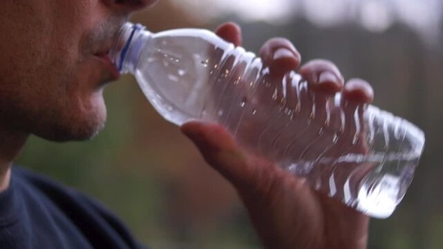 Close-up shot of a man hydrating with water from a disposable, single-serving plastic bottle against a natural, blurred background
