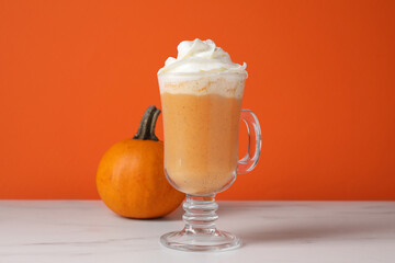 Warm pumpkin latte with whipped cream on white marble table against orange background