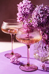 Two champagne glasses and blooming lilac bouquet in soft purple light on pastel surface with delicate shadows
