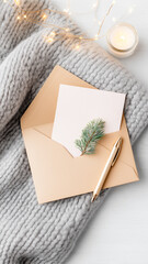 A vertical flat lay photograph featuring a brown kraft paper envelope with a blank card inside, resting on a chunky grey knitted blanket. A gold pen and a small green pine sprig decorate the envelope.