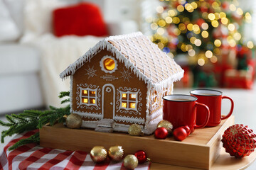 Beautiful gingerbread house, mugs, fir branch and baubles on table against blurred lights indoors, closeup. Christmas atmosphere