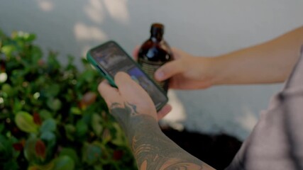 Tattooed young man holding smartphone and open bottled coffee outdoors near green bushes, closeup of hands using mobile app, lifestyle concept for coffee brand, social media and digital marketing
