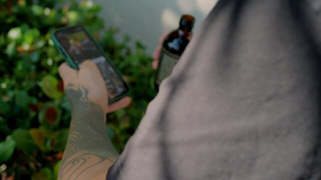 Over‑the‑shoulder view of tattooed young man holding smartphone and bottled coffee outdoors near green bushes, scrolling social media, lifestyle shot ideal for coffee brand and mobile app marketing