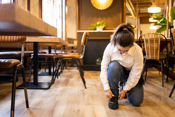 A young woman between 20 and 30 years old with Down syndrome is working as a waitress in a cafeteria.The girl ties the knot of her uniform shoes. Concept of trisomy 21.