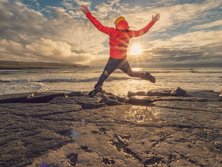 A man in a red jacket stands on a rock overlooking the ocean. The sky is cloudy and the water is...