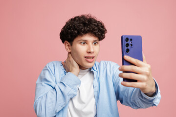 Shocked young man taking smartphone selfie reacting to unexpected news on pink background