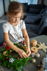 Naklejka premium Little girl finishing her handmade Christmas wreath by tying a string to hang it. Final step of a festive DIY project captured at home in a cozy family setting.