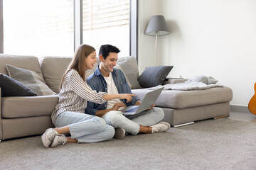 Young 30s couple sitting on floor with laptop in cozy living room, planning vacation, organizing trip, managing household finances, searching design ideas and making orders. Modern digital lifestyle