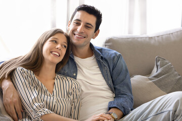 Young 30s beautiful couple in love sitting close on cozy couch, smiling and enjoying peaceful,...