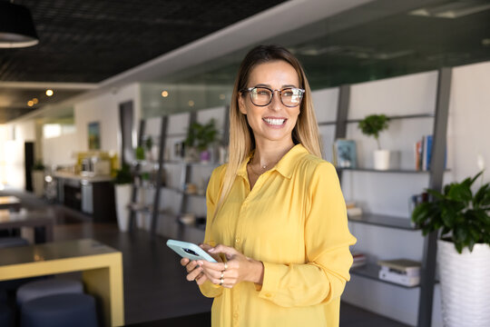Happy beautiful young entrepreneur woman in glasses typing on smartphone in co-working hall, looking away with toothy smile, thinking on online communication, Internet technology for business - Powered by Adobe
