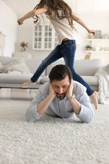 Parenting is hard work. Exhausted upset mid aged dad lying on warm living room floor hug head in despair unable to deal with active energetic small daughter jumping running creating noisy chaos around