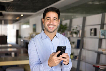 Happy handsome entrepreneur standing with smartphone in office co-working space, holding mobile phone, looking at camera, smiling posing for portrait, using Internet technology for business