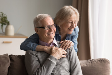 Bonding with laughter and dreams. Loving elderly spouses relax at home together looking away. Happy smiling aged husband enjoy tender hugs of dear senior wife share hopeful ideas joyful future plans