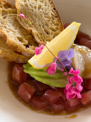 Beautifully decorated tuna tartare dish with avocado and scallop slices, lemon, bread chips and decorated with flowers on a white plate