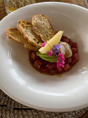 Beautifully decorated tuna tartare dish with avocado and scallop slices, lemon, bread chips and decorated with flowers on a white plate