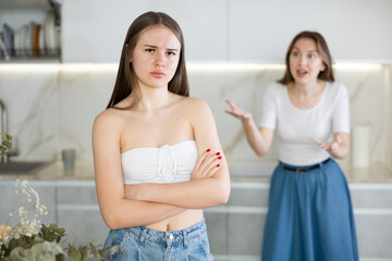 Upset teen girl standing in the kitchen, cross mother scolding her standing behind