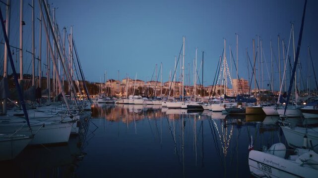 Wide shot of a harbor in a southern French coastal town at dusk. Boats sit still on the water as warm building lights reflect off the surface