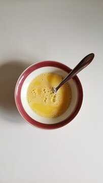 Beaten eggs with bubbles in a small white bowl with a red rim and a metal spoon inside. - Ovos batidos com bolhas em uma pequena tigela branca com borda vermelha e uma colher de metal dentro