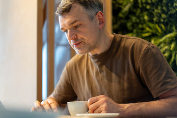 Man in casual brown shirt focuses on work at a laptop while holding a coffee cup in a well-lit cafe
