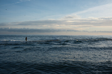 A lonely fisherman stands waist-deep in the tranquil sea, casting his line against the vibrant backdrop of a setting sun. Capturing feelings of peaceful solitude, hope, and the quiet contemplation