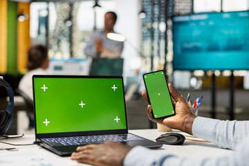 Closeup of black man using smartphone and laptop with blank chroma key display in office workspace. African american corporate professional working with digital devices displaying green screen.
