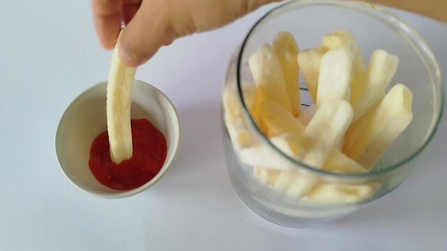 Hand taking a crispy potato stick from an open glass jar and dipping it into a small white bowl of tomato sauce on a clean white background, perfect for snack ads and product demos.