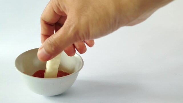 Hand dipping a crispy potato stick into a small white bowl of tomato sauce on a clean white background, ideal for snack promotions and minimalist food visuals.