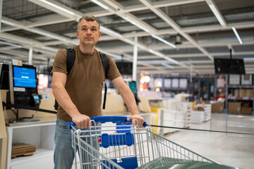 Man wearing a brown shirt pushes a shopping cart in a large supermarket with bright lighting.