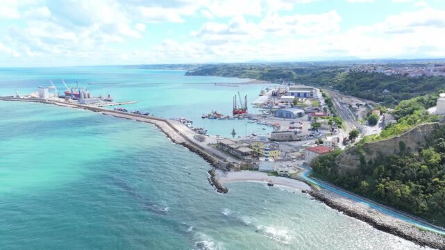 Drone shot of Ortona harbor showing turquoise waters, quiet waves, vessels and port facilities beneath soft white clouds.
