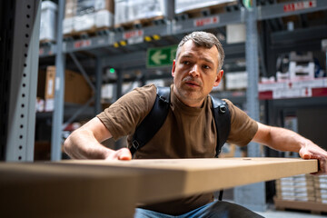 Warehouse worker wearing a brown shirt lifts a cardboard box from a shelf surrounded by industrial shelves and products