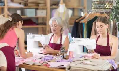 Team of tailors at work, three women in dressmaker office workroom. Two craftsperson designer sews clothes, perform edge finishing stitching seam, assistant attaches pattern to fabric..