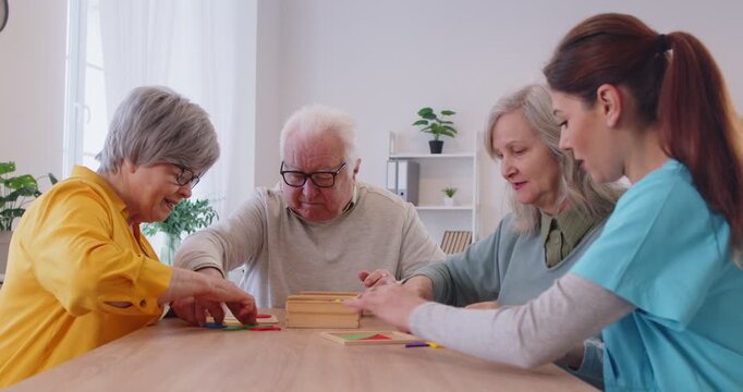Senior people collecting jigsaw of geometric figures sitting at table in nursing home with young friendly nurse. Memory training for dementia prevention and retirement leisure concept. 4k video.
