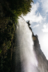 Majestic waterfall cascading down rocky cliffs, surrounded by lush greenery and bright blue sky,...